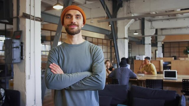 Cool hipster guy smiling at camera with folded hands in office with coworkers