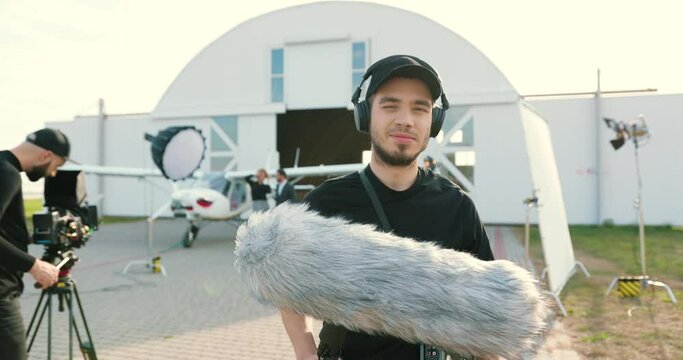 Portrait of happy handsome young Caucasian man in black in headphones standing in foreground outdoor with microphone. Male professional with filming equipment on on set. Film crew on background