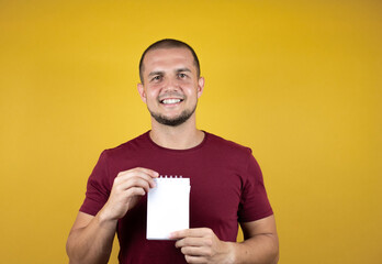 Russian man wearing basic red t-shirt over yellow insolated background smiling and showing blank notebook in her hand