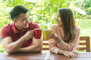 Cheerful young couple holding cup of coffee or tea while sitting on chair in the garden at home. Romantic lover enjoying drink hot mug mocha on the cup glass together in the morning.