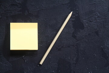 On a black textured rough background, a part of a yellow paper sticker peeks out and a wooden pencil lies on top.