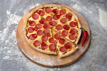 Whole Spicy Italian Pepperoni Pizza with salami slices, fresh chilli pepper and mozzarella cheese served on wooden plate served with culinary flour and spices on gray concrete background.

