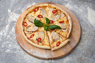 Whole spicy Pizza with sliced pork and chicken meat, mozzarella cheese, chilli pepper slices served with fresh basil on top on wooden plate served with culinary flour and spices on concrete background