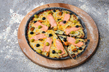 Whole seafood black dough Pizza on wooden plate served with culinary flour and spices on gray concrete background. Fast food Dish With salmon slices, green and black olives and mussels
