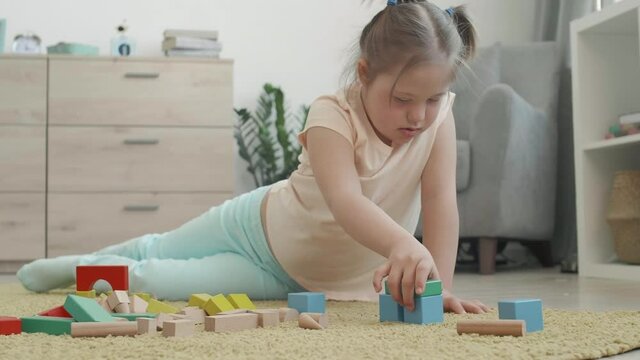 Lockdown of Caucasian adorable handicapped schoolgirl sitting on carpet in living room and playing with tinker toys