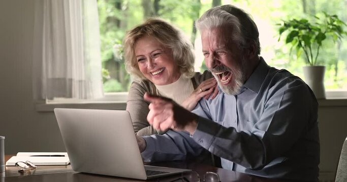 Elderly spouses advanced users of modern tech sit at table looking at laptop screen spend free time funny using device and internet web page. Mature couple laughing watch videos online feels carefree