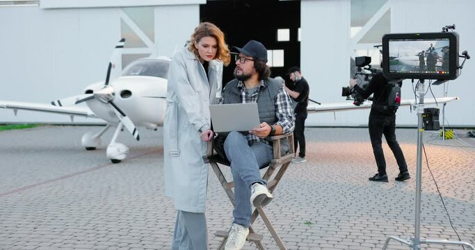 Handsome male film director in glasses and cap sitting in chair on backstage and typing on laptop outdoor. Caucasian beautiful woman talking to man who is tapping on computer on set. Backstage concept
