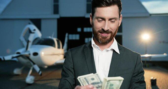 Close up portrait of handsome Caucasian young businessman counting money while standing outdoors in spotlight. Joyful male with many dollars in evening. Luxury life. Money concept