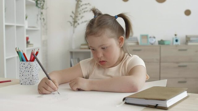 Medium shot of cute handicapped schoolgirl sitting at desk at home and drawing using colour pencils