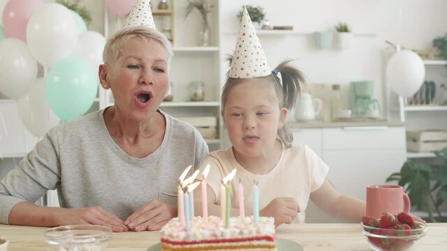 Lockdown of joyful old woman and her cute granddaughter with down syndrome sitting at kitchen table and blowing out candles on birthday cake