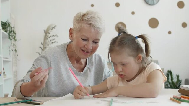 Medium shot of blond-haired senior woman and schoolgirl with down syndrome sitting together at desk at home and drawing using colour pencils