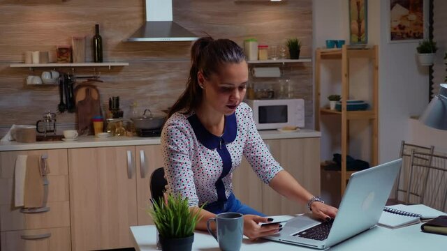 Hispanic woman using credit card in home kitchen to make online payment. Employee making paymant transaction from home on digital notebook conected to internet working overtime for job