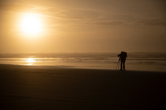 A distant backside view of a photographer on the beach at sunset 

