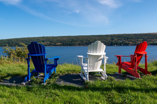 Three Adirondack chairs one red, blue and white sits on the edge of the ocean on a sunny summer's day. There's a tree covered mountain in the distance.  There's a view from the relaxing wooden chairs.