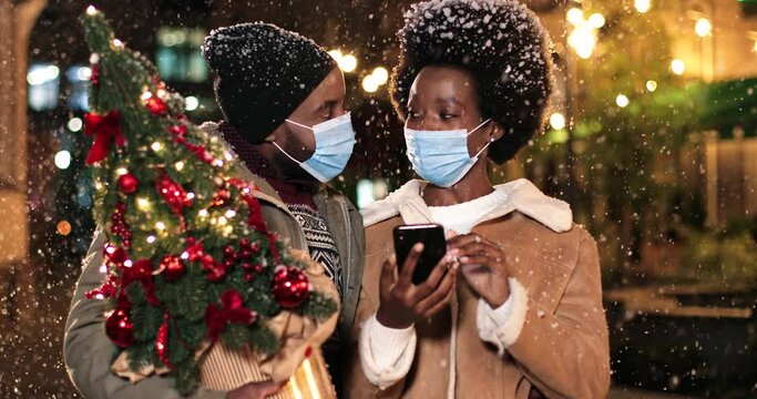 Close Up Of Cheerful Family Standing In Snowy City And Talking While Snowing. Young African American Happy Female Tapping On Smartphone Outdoors With Man With Little Christmas Tree. Winter Concept