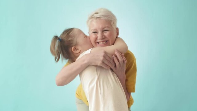 Medium shot of Caucasian elderly woman with short haircut and teenage girl with down syndrome standing against light blue background, looking at camera, hugging and smiling