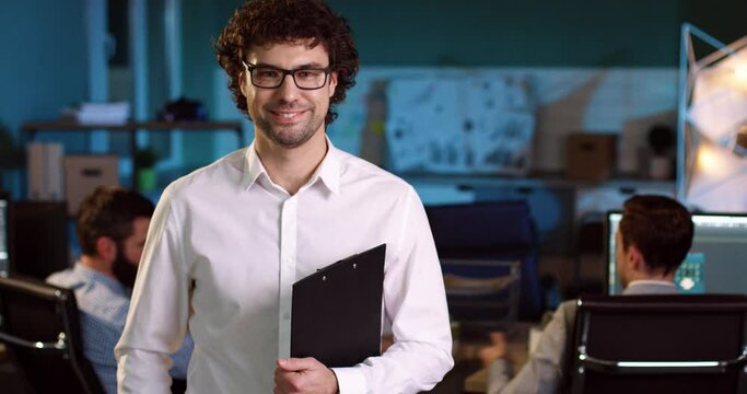 Close up portrait of adult businessman in white shirt with glasses standing in the middle of the office with folder in his hands and smiling.