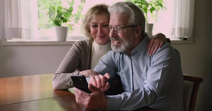 Elderly wife hug hoary husband in glasses sit together at table at home relaxing using smartphone, having fun on internet, read media news, spend free time buy choose goods and services online concept