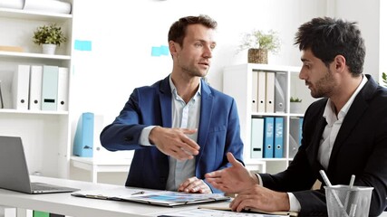 Slow motion medium of two diverse male colleagues sitting at desk in bright office talking and looking at business papers, shaking hands saying goodbye after productive workday