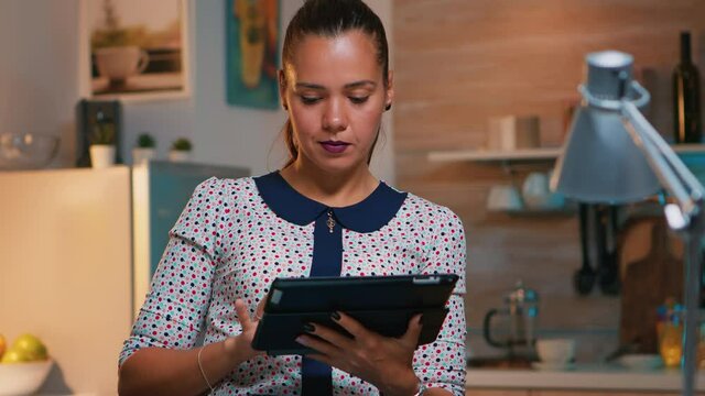 Businesswoman taking break using tablet late at night sitting in modern kitchen. Busy focused employee using modern technology network wireless doing overtime writing, searching.