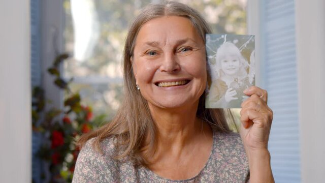 Attractive retired woman with grey hair holding her photo as little girl