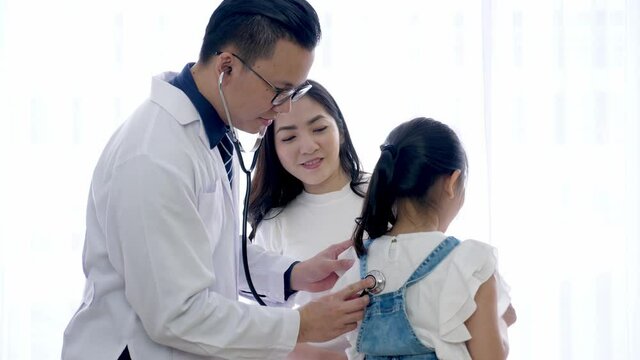 Pediatrician and kid concept. Asian male doctor put stethoscope on the back of female child patient to listen to lungs functional while the girl breath in and out, and the mother standing besides.