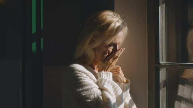 Sick senior woman coughing hard standing near window at home