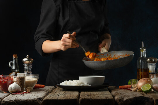 Professional chef pours by ladle chicken curry from pan wok to white bowl. Backstage of serving traditional Indian curry with boiled rice on black background. Concept of cooking tasty hot meal.