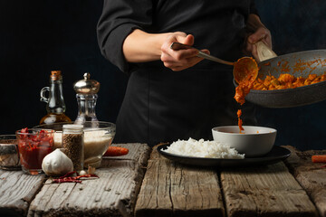 Close-up view of chef pours by ladle chicken curry from pan wok to white bowl. Backstage of serving traditional Indian curry with rice on wooden table with ingredients background. Frozen motion.