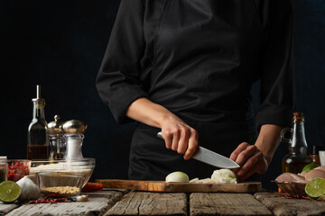 The professional chef in black uniform cuts with knife whole white onion on chopped wooden board at rustic table with ingredients for cooking background. Backstage of preparing meal with vegetables.