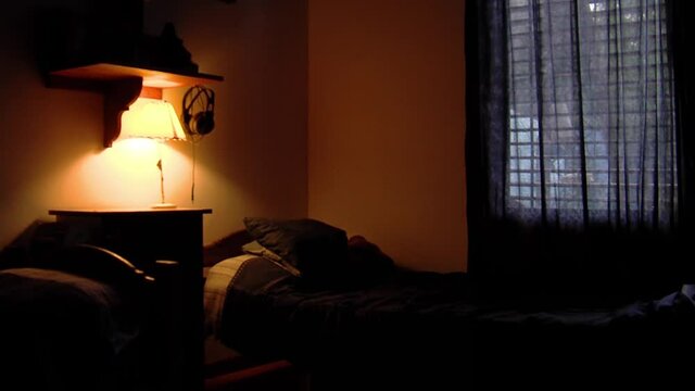 Lamp on a Night Table next to a Bed Spreading Dimmed Yellow Light in the Room. Zoom Out.