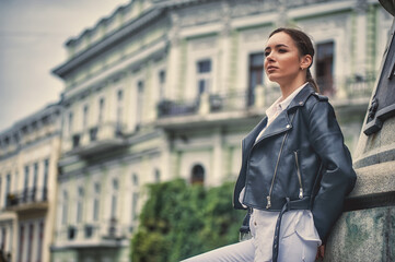 Portrait of a young beautiful woman in a leather jacket and light trousers on the street of a European city