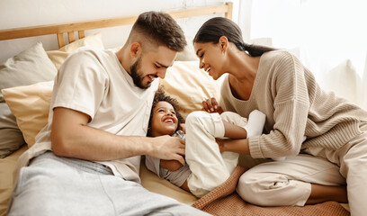 happy diverse family mom, dad and child  laughing, playing and tickles   in bed   at home.