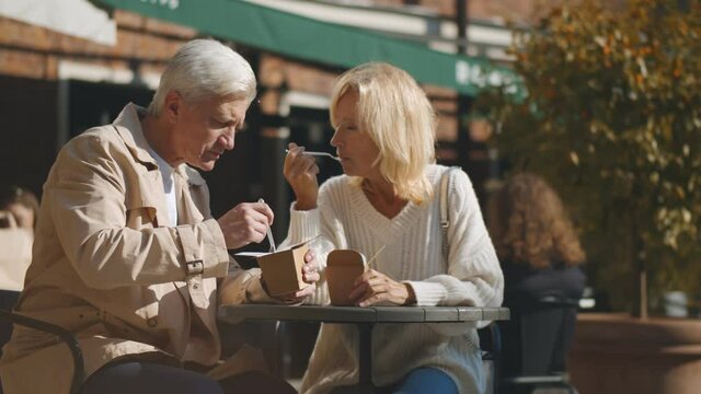 Aged couple sitting at small outdoor cafe table and enjoying takeaway lunch