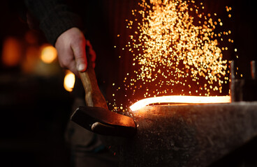 The blacksmith hits the red-hot workpiece in the forge with a hammer and glowing sparks fly in all...