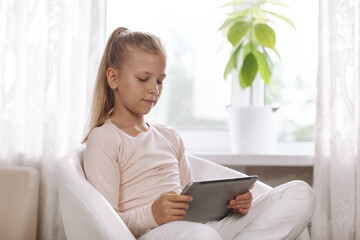 Beautiful teenage girl sitting in white armchair in white room doing homework using tablet, online education, distance learning, technology concept