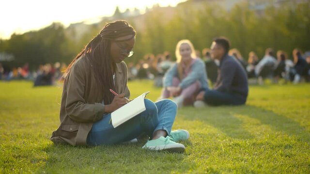 Afro female student writing in exercise book and doing homework in summer park