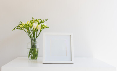 Close up of white freesia flowers in glass vase next to blank square picture frame on white table against wall (selective focus)
