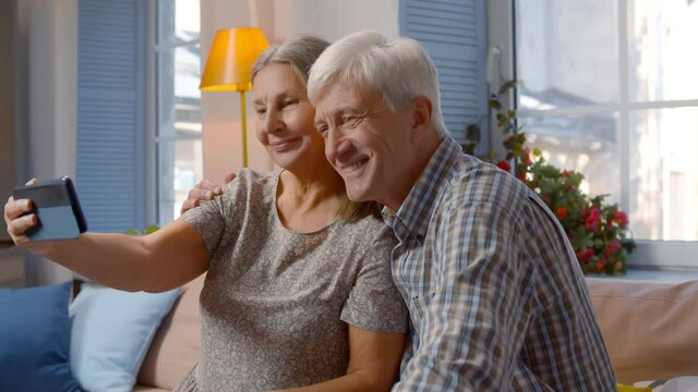 Excited elderly couple sitting on couch making selfies on cellphone