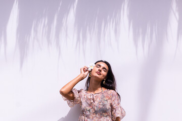 Happy smiling laugh caucasian long hair woman in dress stands on white wall background, holding thai asian flower. Beautiful shadow falls behind. Sunny day, outdoor shot. 