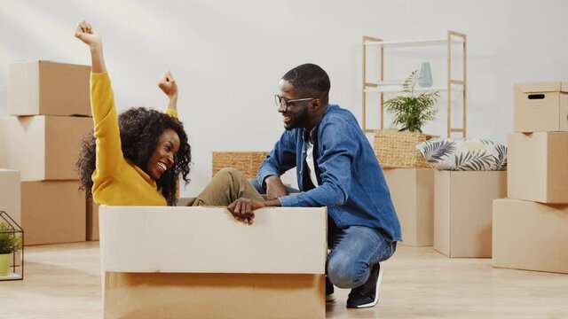 Happy young couple moved to new apartment. Cheerful woman sitting in empty cardboard box. Funny man and woman raise their hands up.