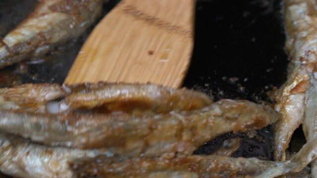 Cooking frying and stirring capelin fish fried in iron pan. Close-up view chief-cooker stirs roasted caplin fish with wooden spatula. Grilled fish - traditional Asian dish as an appetizer or garnish