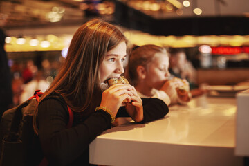 Beautiful schoolgirl eating hamburger sitting in a cafe indoors. Happy child eating junk food in a restaurant. Side view