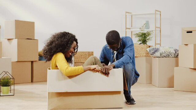 Happy young couple moving to new apartment. Cheerful adult girl sitting in cardboard box on floor. Funny man and woman joking and laughing.
