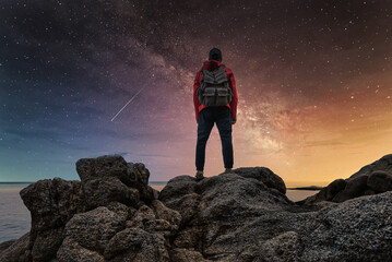 A man in a red jacket with a backpack on his back stands on the rocks by the sea at night and looks at the starry sky and the milky way over sea