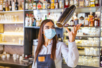 Beautiful female bartender with protective face mask tosses cocktails shaker into the air and...