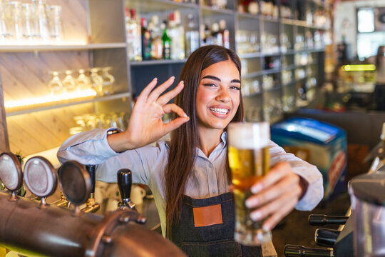Beautiful smiling female Bartender showing ok sign serving a draft beer at the bar counter , shelves full of bottles with alcohol on the background