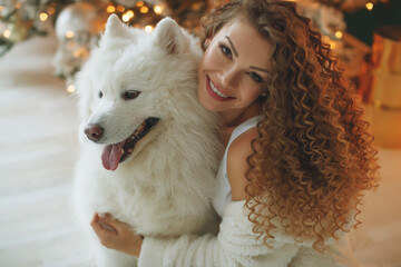 Beautiful woman sits on a white blanket with a dog hugs, against the background of a Christmas tree fireplace with candles in a decorated room. Happy New Year and Merry Christmas. High quality photo.