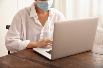 Fototapeta premium Junge Frau mit weißer Bluse arbeitet am Laptop und trägt einen Mundschutz.