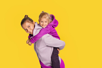 Smiling blonde kid girl and woman in stylish jumpsuits with hood posing over yellow background. Cute daughter 4-5 year old hugging her mother. Mock up copy space lifestyle concept.